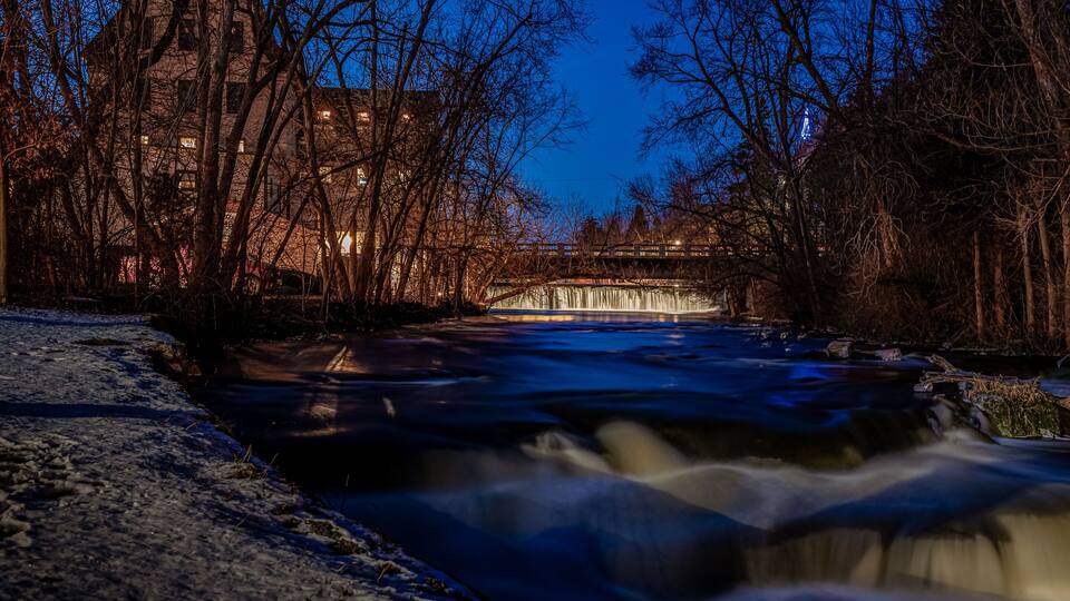 A river flowing through a city at night with buildings and a bridge in the background.