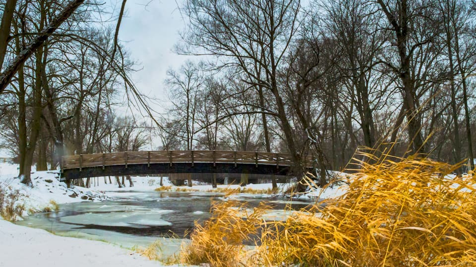 A wooden bridge over a partially frozen river, surrounded by snow-covered ground and leafless trees.