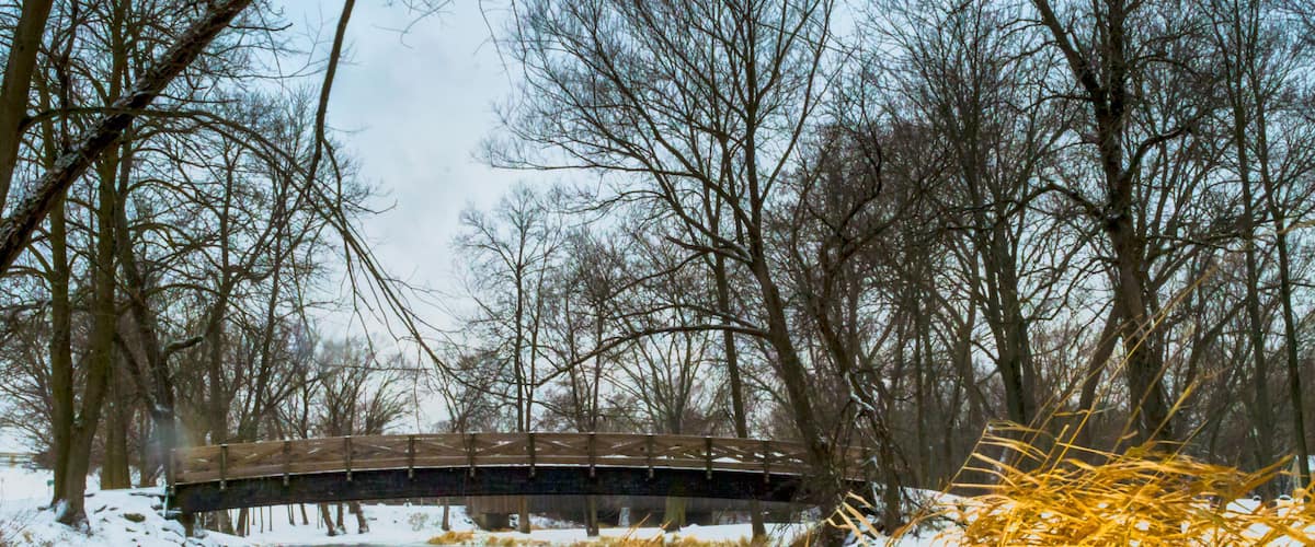 A wooden bridge over a partially frozen river, surrounded by snow-covered ground and leafless trees.