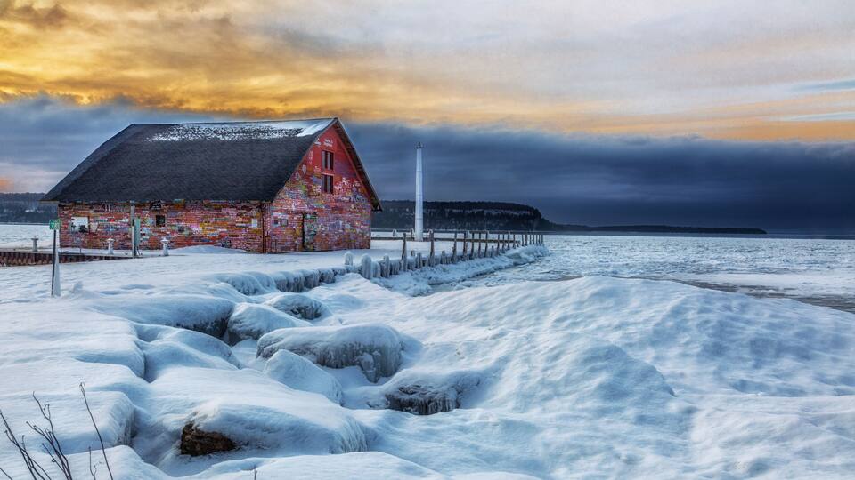 A red wooden building with snow-covered ground and a lighthouse in the background.