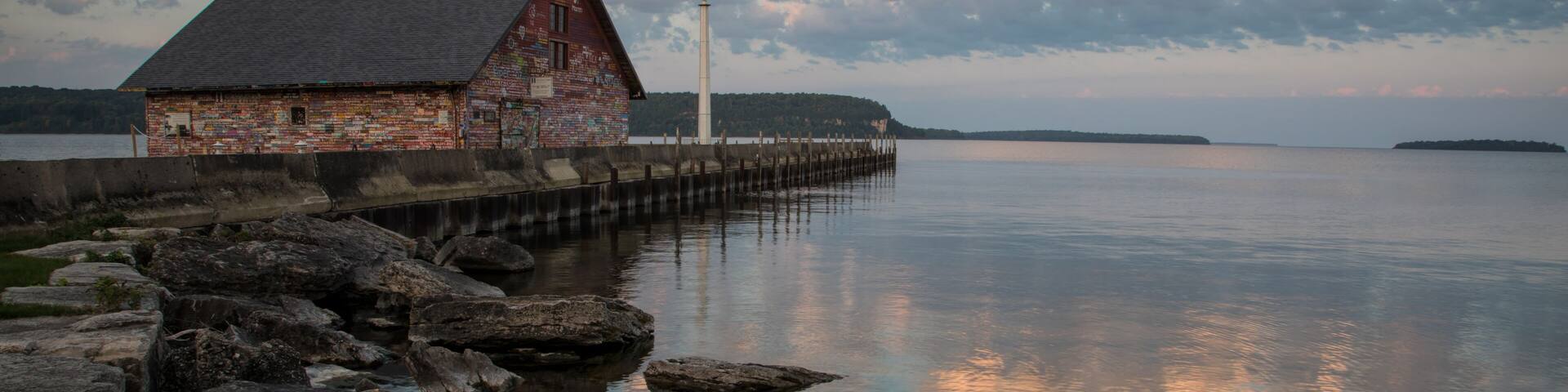 A rustic wooden building by a calm lake with a pier extending into the water, surrounded by large rocks.