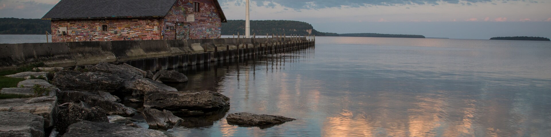 Un edificio rústico de madera junto a un lago tranquilo con un muelle que se adentra en el agua, rodeado de grandes rocas.
