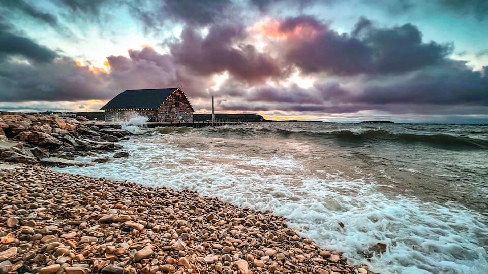 A rocky beach with waves crashing onto the shore, a wooden pier, and a building with lights.