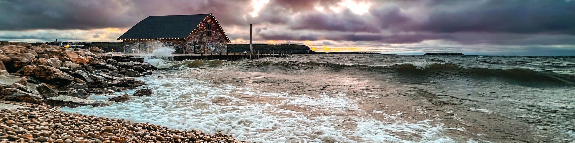 A rocky beach with waves crashing onto the shore, a wooden pier, and a building with lights.