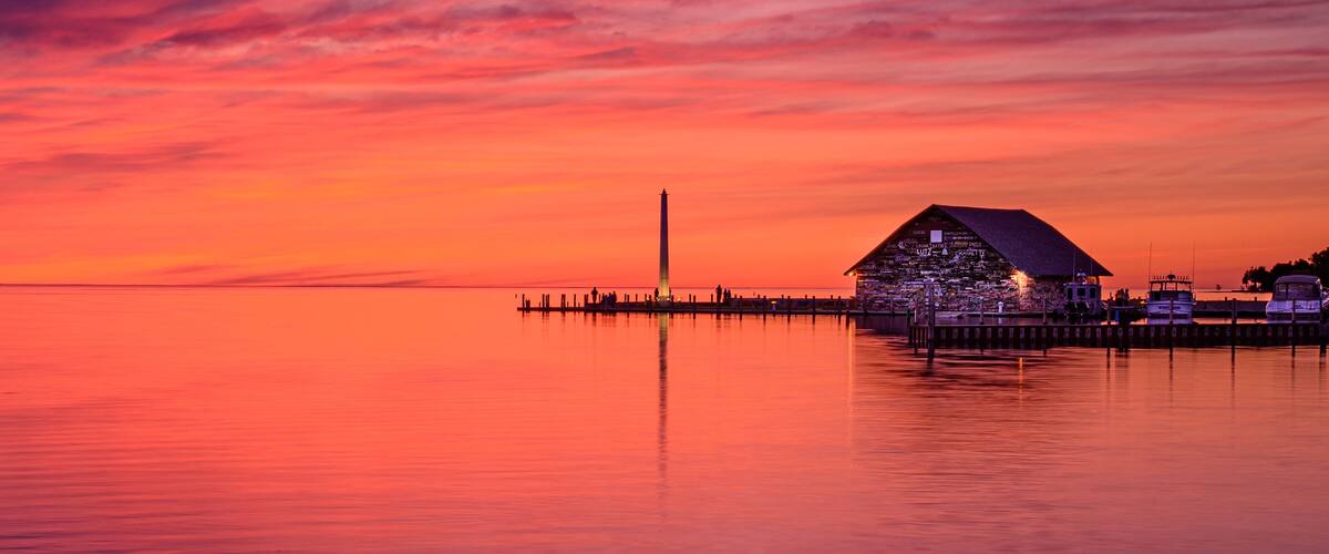 A sunset over a calm body of water with a dock and a building with a pyramid-shaped roof.