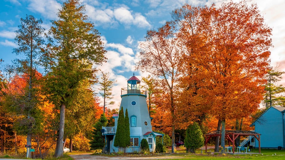 A lighthouse with a red roof, surrounded by autumn-colored trees and a small building with a blue roof.