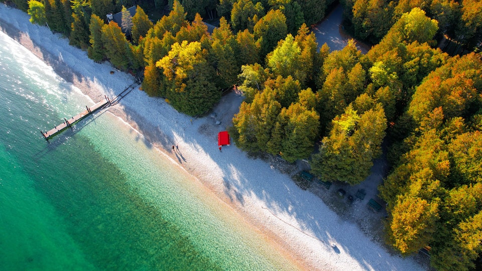An aerial view of a beach with clear turquoise waters, a wooden pier, and a red structure on the shore.