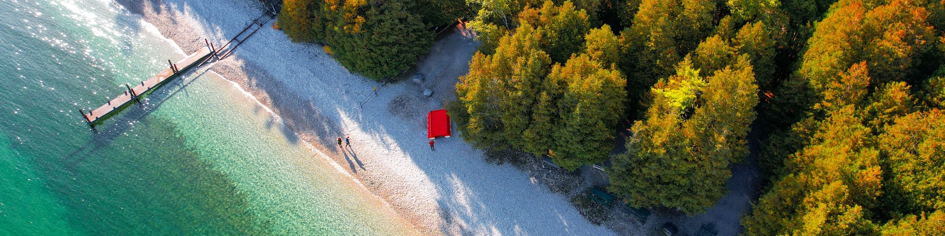 Eine Vogelperspektive eines Strandes mit klarem türkisblauem Wasser, einem hölzernen Steg und einem roten Gebäude am Ufer.
