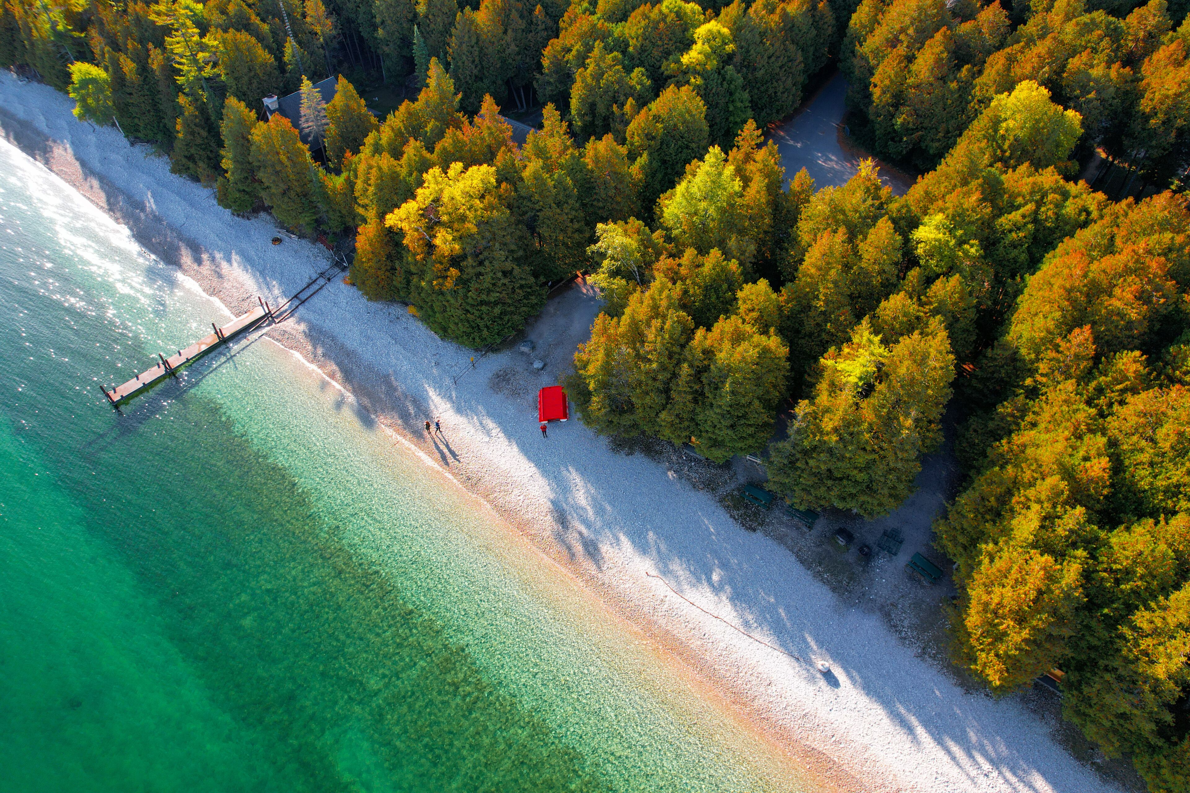 An aerial view of a beach with clear turquoise waters, a wooden pier, and a red structure on the shore.