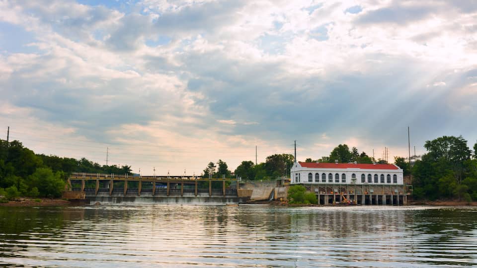 A dam with a bridge, a white building with a red roof, and a cloudy sky over a body of water.