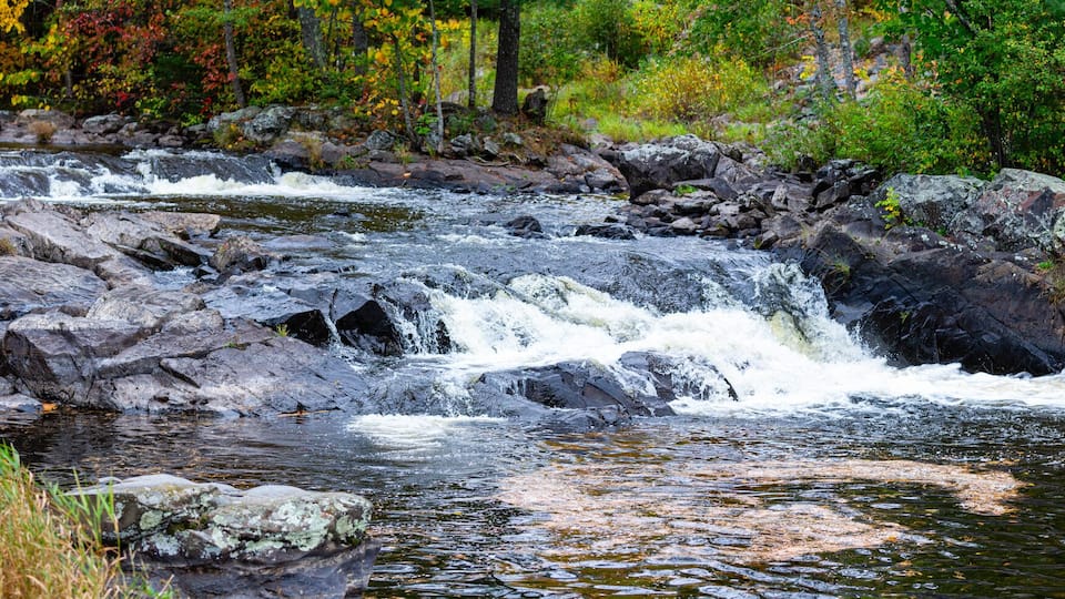 A rocky stream with water flowing over rocks, surrounded by autumn foliage.