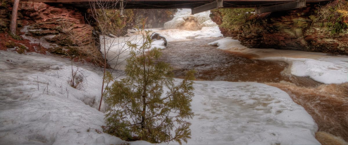 A wooden bridge over a river with snow on the ground and surrounding trees.