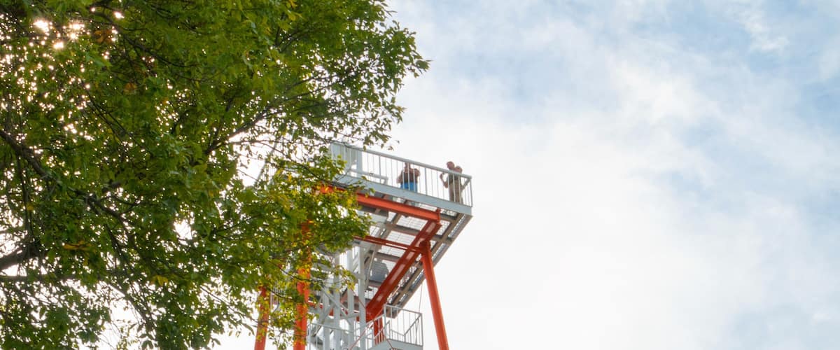 A tall red and white metal tower with stairs and platforms, surrounded by trees and a grassy area.