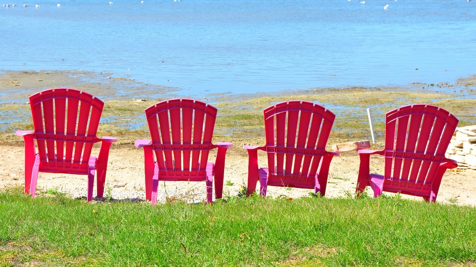 Four red Adirondack chairs facing a beach with clear blue water and a grassy foreground.