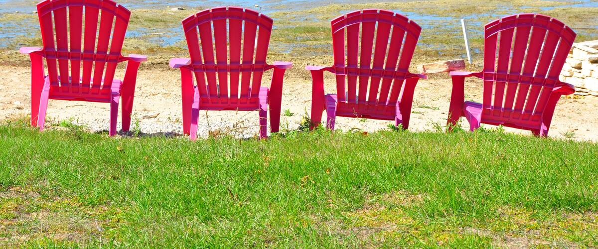 Four red Adirondack chairs facing a beach with clear blue water and a grassy foreground.