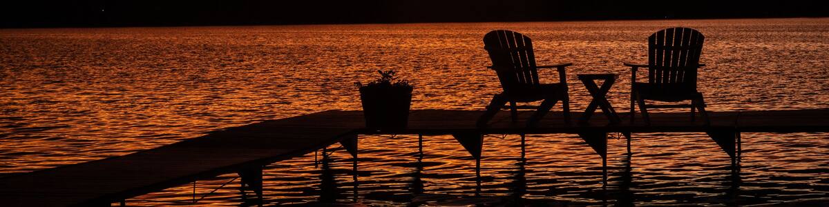 Un coucher de soleil sur un lac, avec deux chaises Adirondack et une table sur un quai.