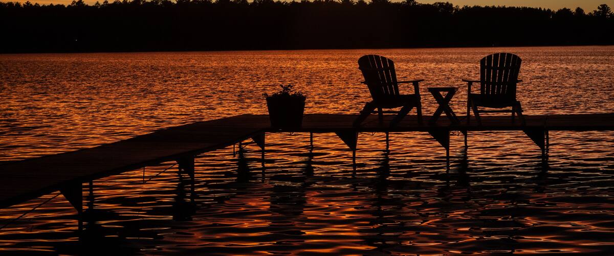 Un coucher de soleil sur un lac, avec deux chaises Adirondack et une table sur un quai.