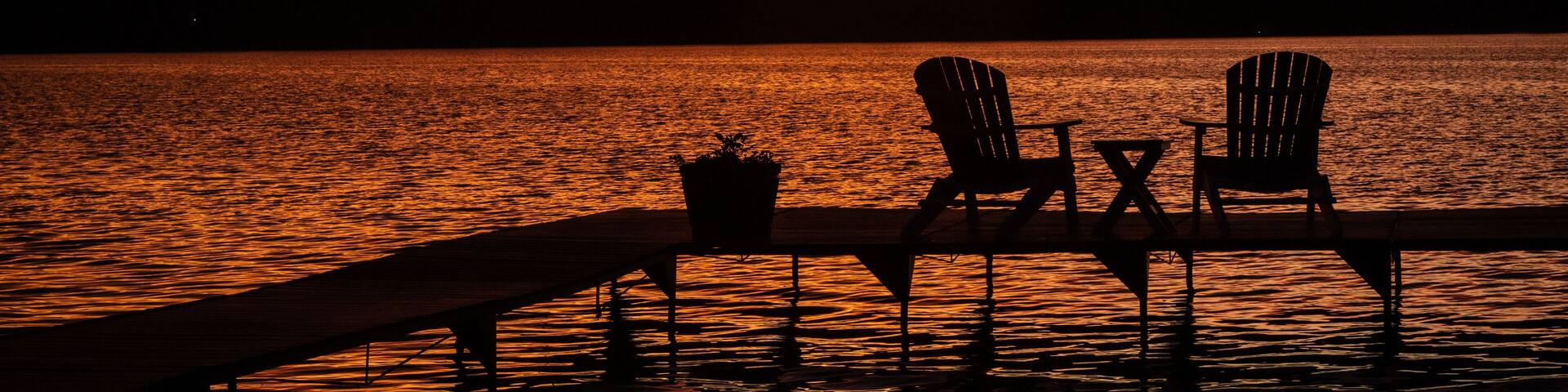 A sunset over a lake with two Adirondack chairs and a table on a dock.