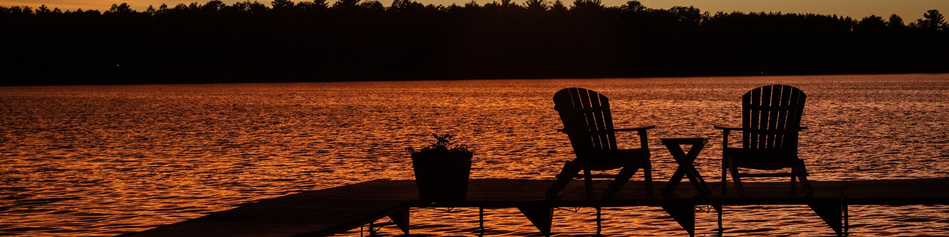 Atardecer sobre un lago con dos sillas Adirondack y una mesa en un muelle.