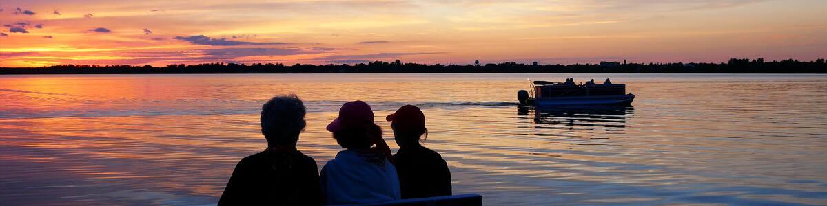 Silhouette of 3 senior women enjoying a colorful sunset on a bench at a beautiful lake as a pontoon passes by.