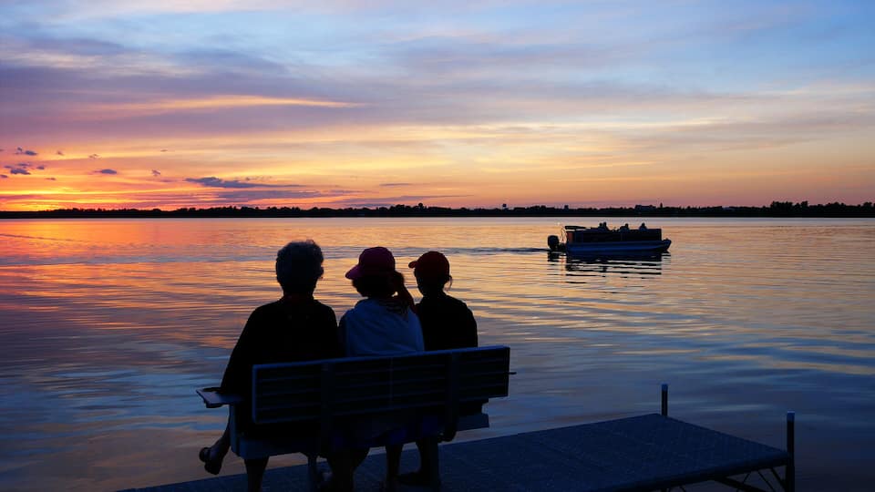 A sunset over a calm lake with a boat in the distance and a bench overlooking the water.