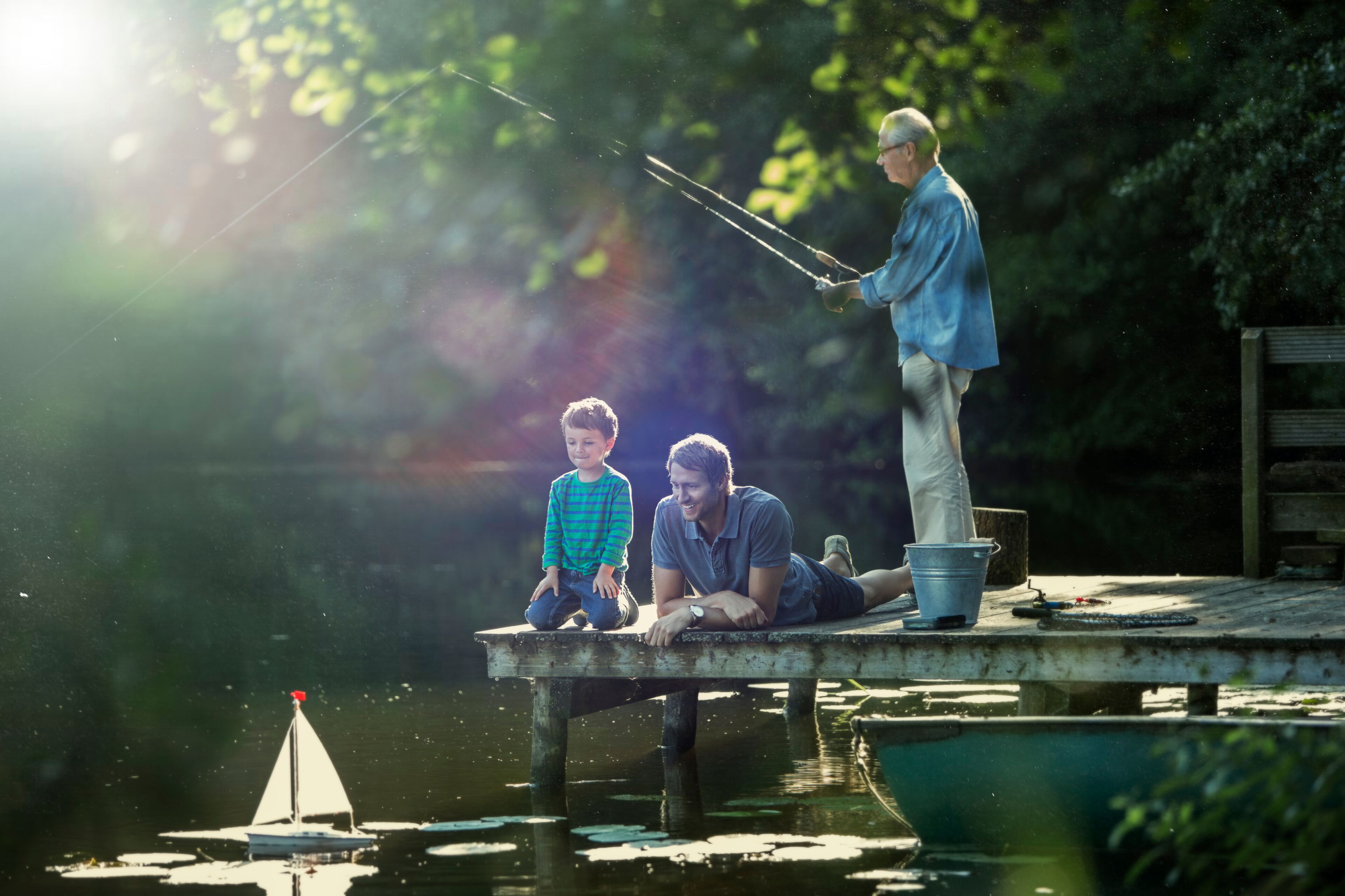 Boy fishing playing with toy sailboat with father grandfather at lake