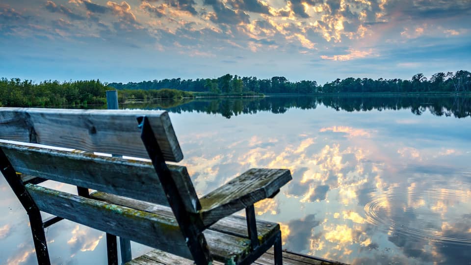 A wooden bench by a calm lake with a reflection of the sky and trees.