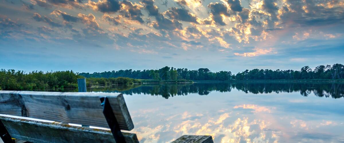 A wooden bench by a calm lake with a reflection of the sky and trees.