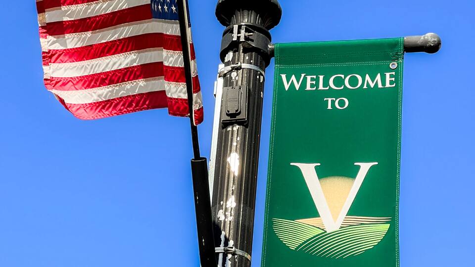 An American flag, a street lamp, and a banner with 'WELCOME TO VIRQUA' and 'ABUNDANT LIFE' against a clear blue sky.