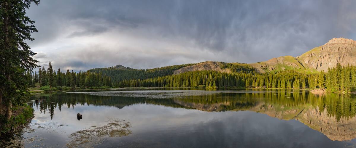 A calm lake surrounded by dense forest and mountains under a cloudy sky.