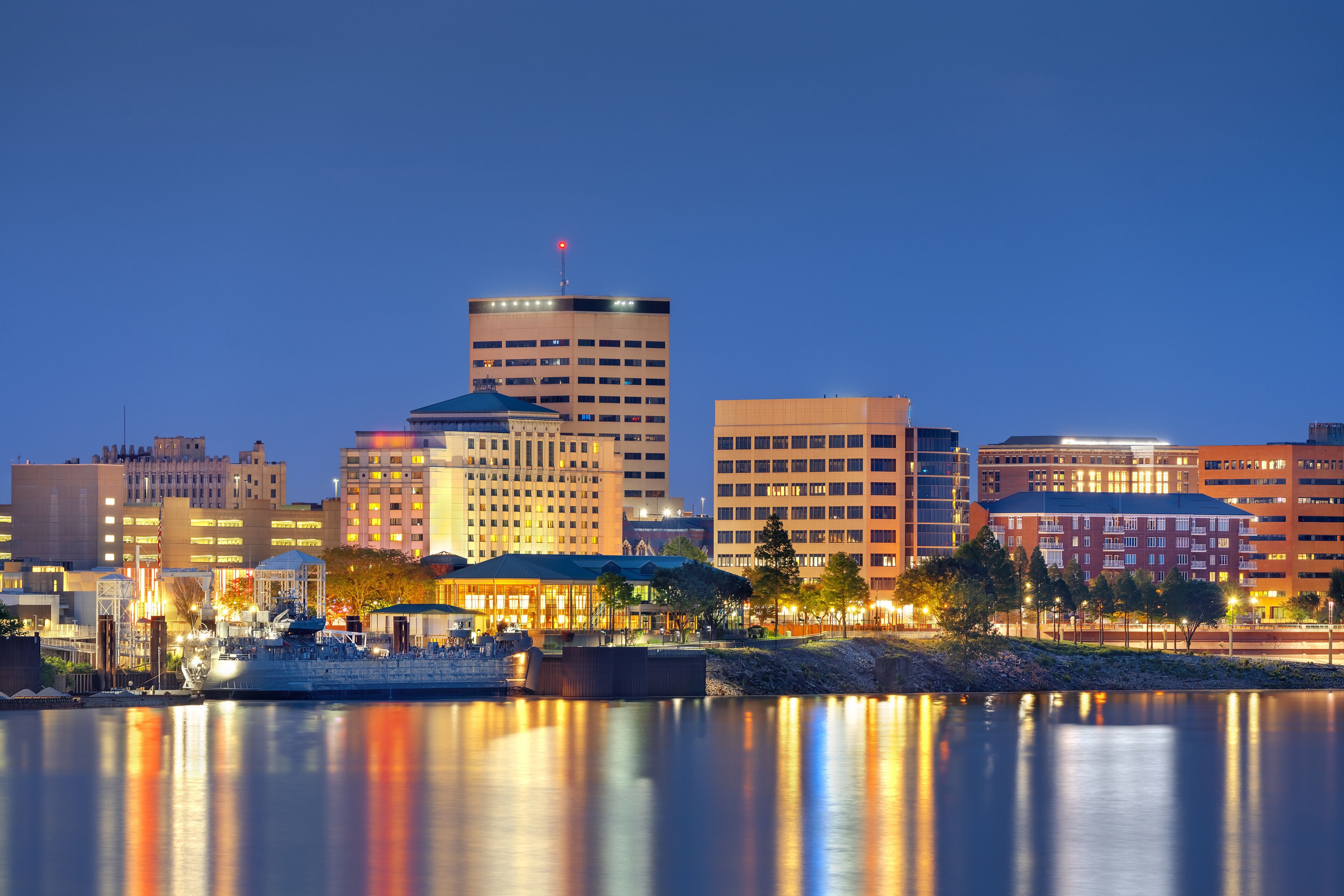 A city skyline at dusk with illuminated buildings reflecting on the water.