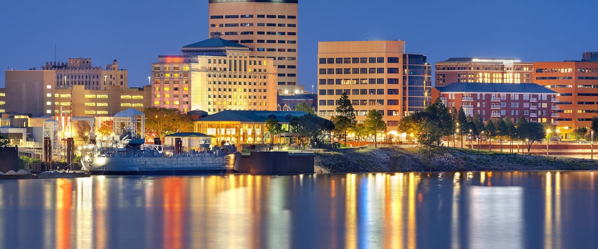 A city skyline at dusk with illuminated buildings reflecting on the water.