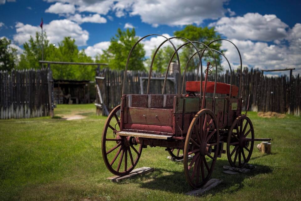 Un antiguo carro cubierto en un campo de hierba, con una cerca de madera y árboles al fondo.
