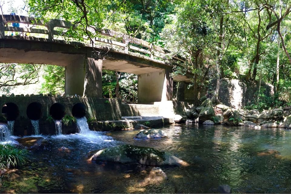 A stone bridge over a clear stream with water flowing over rocks, surrounded by lush greenery.