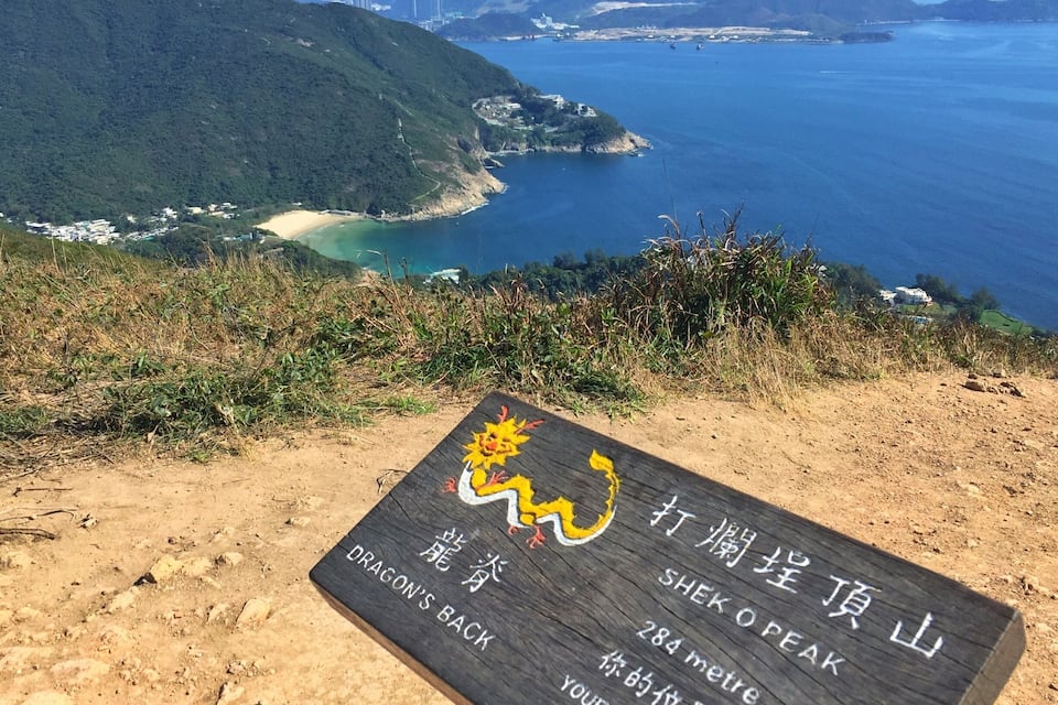 A sign on a hillside with a scenic view of a coastal area, including mountains, a beach, and a body of water.