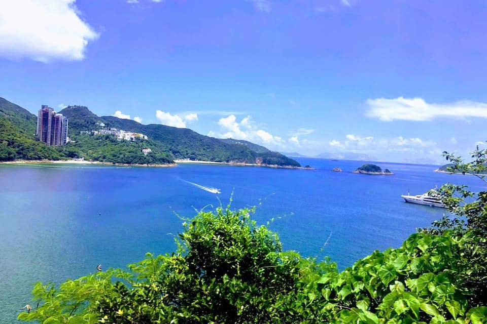 A coastal landscape with a clear blue sea, a yacht, and a building on a hill.