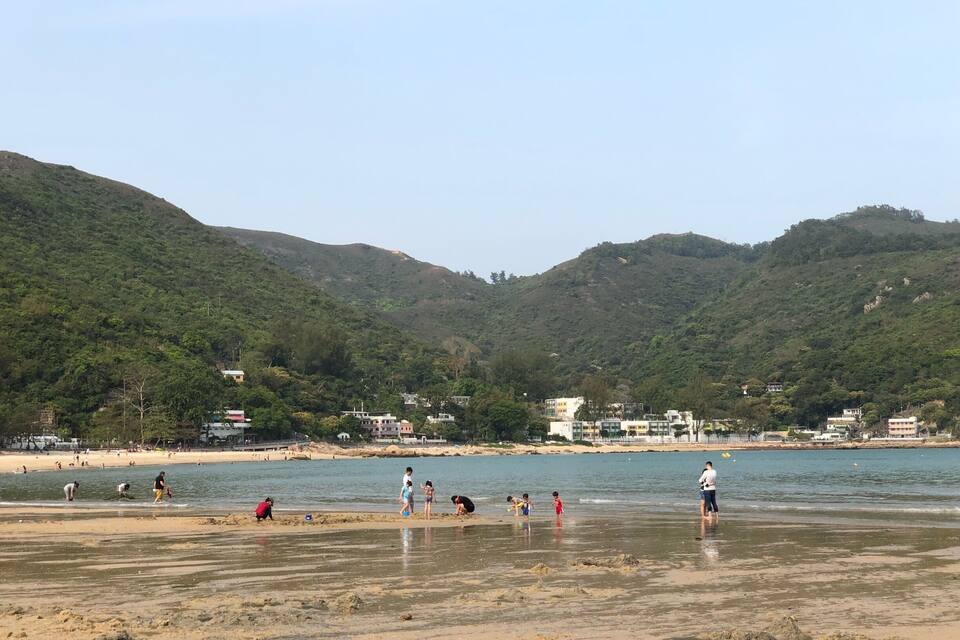 Uma praia com pessoas brincando em águas rasas, rodeada por colinas verdes e edifícios.