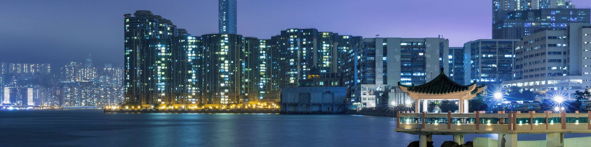 A nighttime cityscape with illuminated high-rise buildings and a traditional pavilion by the waterfront.