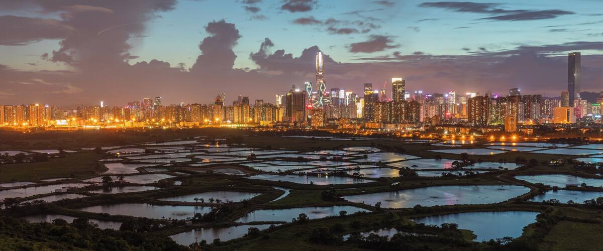 A city skyline at dusk with illuminated buildings and a network of water bodies in the foreground.