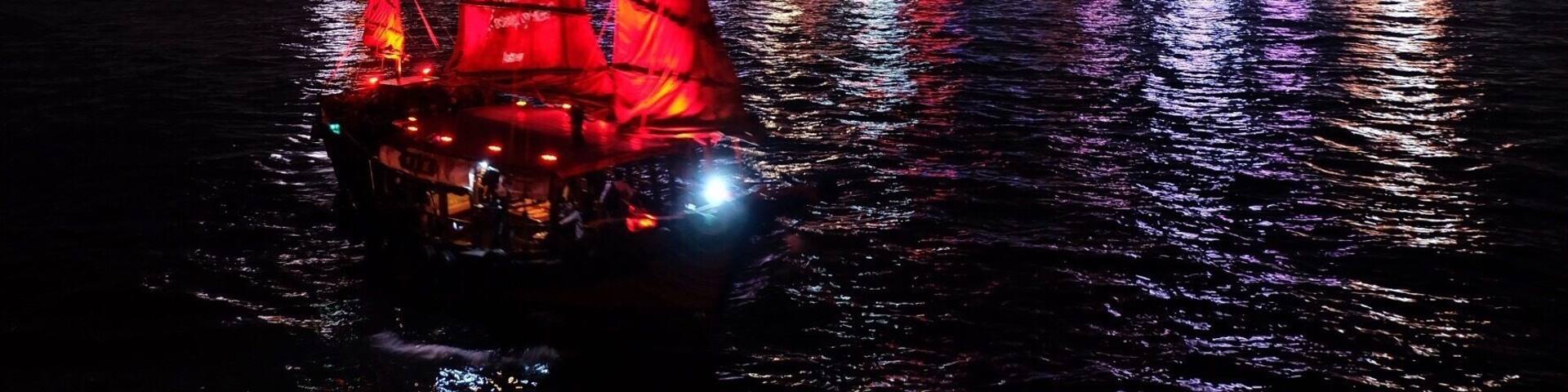 A sailboat with red sails on a body of water with a city skyline in the background.