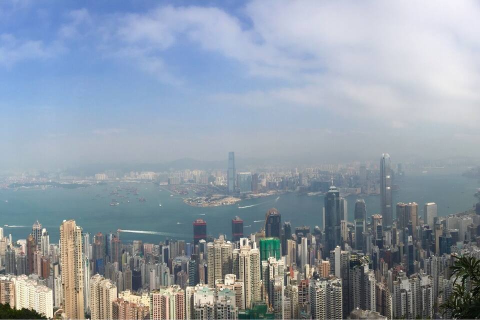 A cityscape with high-rise buildings, a body of water, and distant mountains under a partly cloudy sky.
