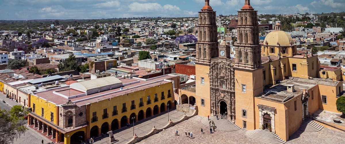 A historic church with two bell towers and a large dome, surrounded by a plaza and urban buildings.