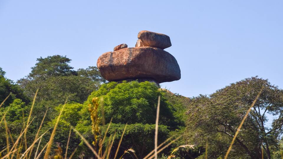 A large rock formation balanced on top of another rock, surrounded by dense green foliage and trees.
