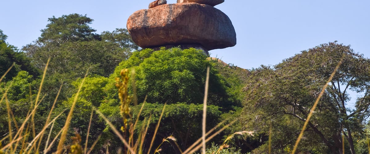 A large rock formation balanced on top of another rock, surrounded by dense green foliage and trees.