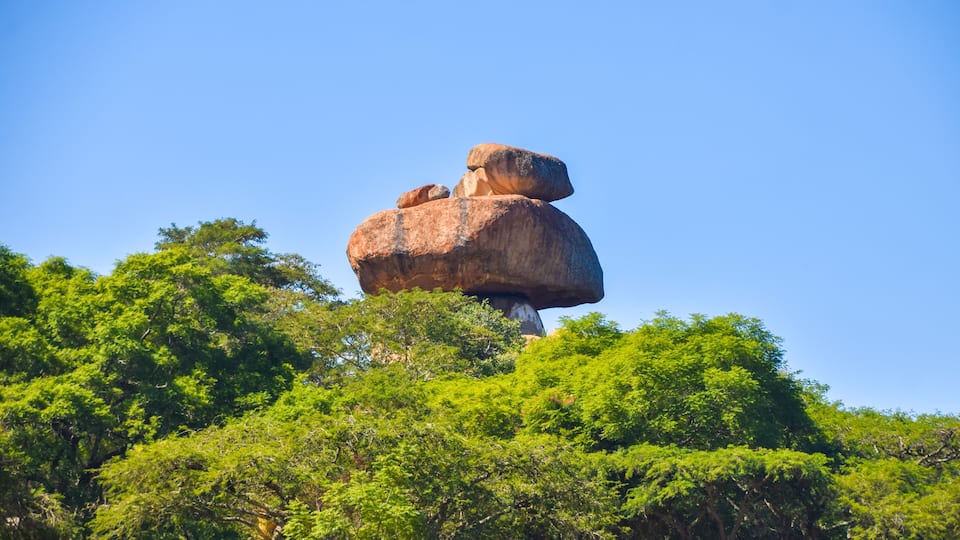 A large rock formation balanced on top of another rock, surrounded by dense green foliage.