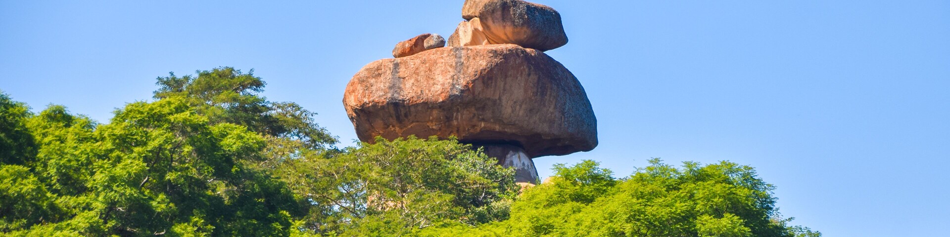 Uma grande formação rochosa equilibrada sobre outra rocha, rodeada por vegetação verde densa.
