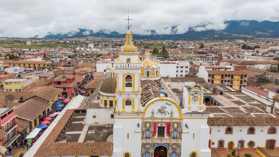 A white church with yellow domes and a bell tower in a town with colorful buildings and mountains in the background.