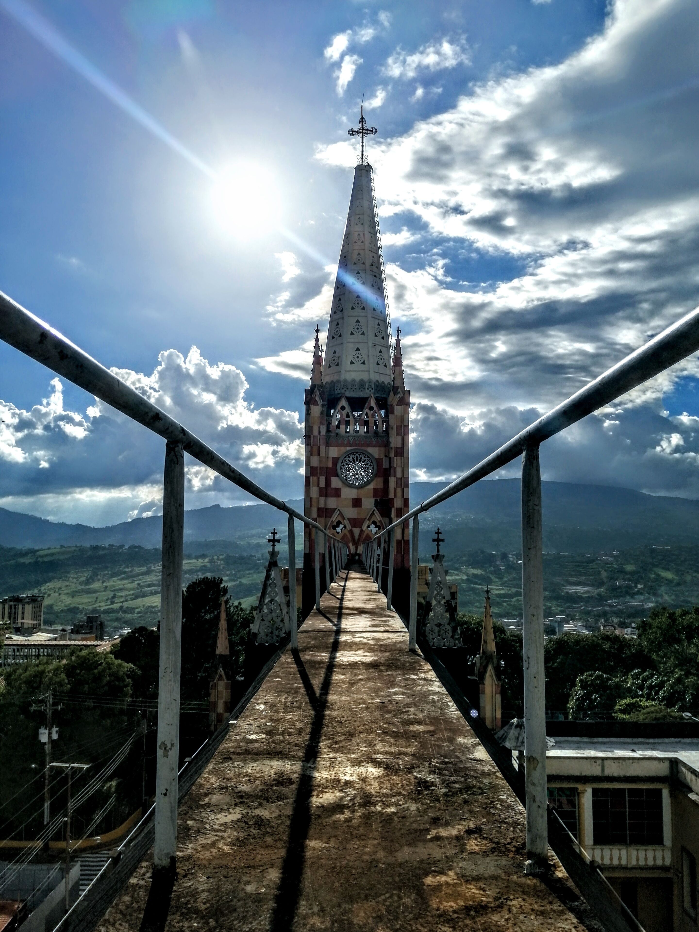 A bridge leading to a tower with a clock and crosses, surrounded by a mountainous landscape.