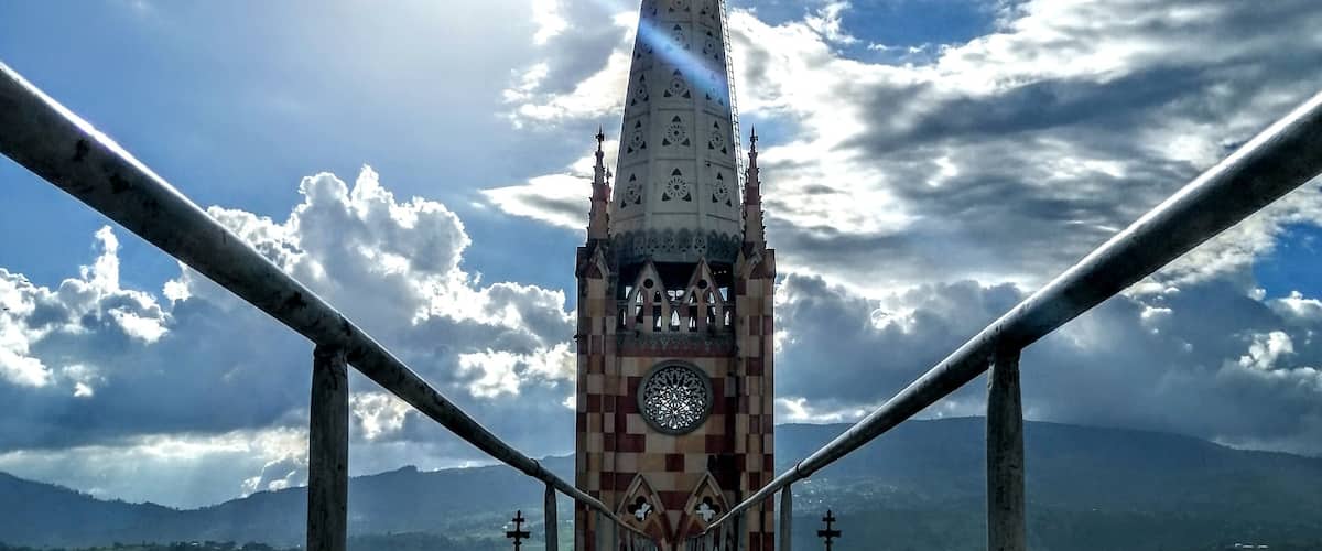 A bridge leading to a tower with a clock and crosses, surrounded by a mountainous landscape.
