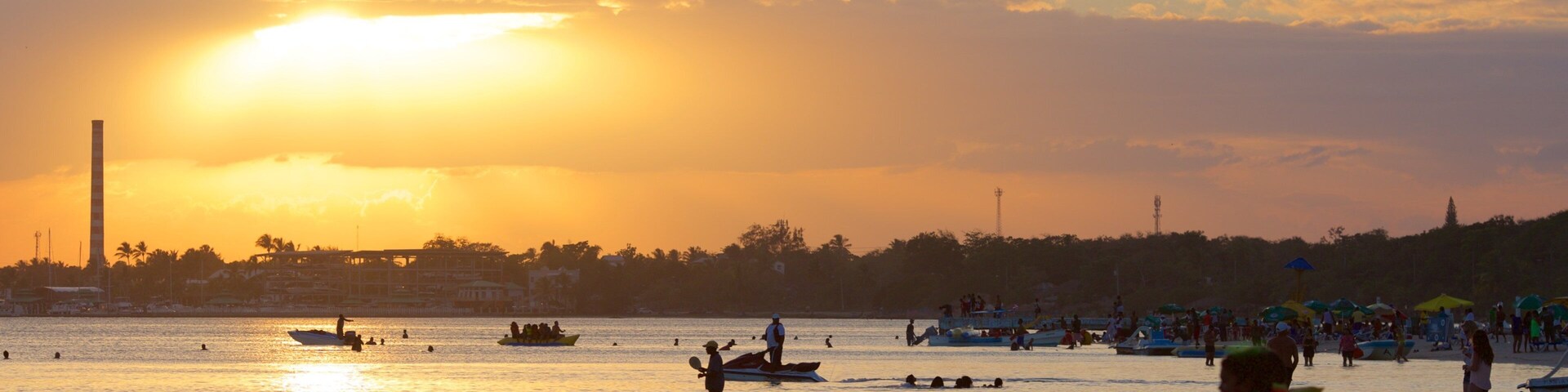 Pôr do sol sobre a água, com barcos e pessoas na margem.